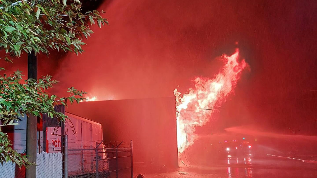 A dramatic night scene showing a large fire burning at the side of a building, with powerful streams of water spraying toward it and bright orange-red flames reflecting across a wet street and puddle.